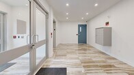 This 220 square foot space serves as a modern and efficient entryway, likely for a multi-unit residential or commercial building. The area boasts a sleek, contemporary design with a focus on functionality. The flooring is a standout feature, showcasing large-format tiles with a luxurious marble-like pattern in warm, neutral tones that add a touch of elegance to the space.

The entryway is well-lit with recessed LED lighting fixtures evenly spaced across the crisp white ceiling, ensuring ample illumination throughout. The walls are finished in a clean, bright white, contributing to the overall sense of spaciousness and modernity.

Key features include a glass entry door system with metal framing, providing natural light and visibility. A blue-gray security door adds a pop of color while maintaining the sleek aesthetic. The space is equipped with essential amenities such as a wall-mounted mailbox system, likely for resident use, and what appears to be an elevator, enhancing accessibility.

Safety features are evident, with visible fire alarm pull stations and emergency signage. The inclusion of a black entrance mat at the doorway demonstrates attention to cleanliness and safety. Overall, this entryway efficiently combines style, security, and practicality in a compact yet welcoming space.