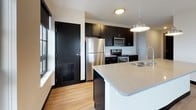 This 2-bedroom, 2-bathroom unit showcases a contemporary design defined by a striking contrast between dark cabinetry, black door frames, and light hardwood flooring that runs throughout the space. The kitchen is a standout feature, equipped with dark espresso cabinetry, stainless steel appliances including a refrigerator, range, and microwave, and a white subway tile backsplash. A spacious island with a light quartz countertop and a deep double sink provides ample workspace and casual seating. A pendant light fixture adds a polished finishing touch above the island.

Both bedrooms are generously sized, each featuring large grid-pane windows with black frames that invite abundant natural light and offer views of the surrounding neighborhood. The light-toned walls and hardwood floors create a clean, airy atmosphere in each room.

The two bathrooms differ in style, offering variety in design. One bathroom features a sleek walk-in shower enclosed in glass, with floor-to-ceiling dark slate-toned tile, a rain showerhead, and a vanity with a dark countertop and blue cabinetry. The second bathroom includes a full tub-shower combination with chrome fixtures and a clean, white surround. Together, the spaces present a cohesive, modern aesthetic throughout.