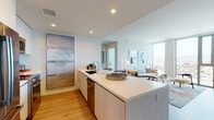 This contemporary high-rise residence is defined by clean lines, a neutral palette, and an abundance of natural light. The open-concept layout connects the kitchen and living area seamlessly, creating an inviting and functional space. The kitchen is outfitted with sleek flat-front cabinetry, a large center island with bar seating, quartz countertops, and a full suite of stainless steel appliances, including a French door refrigerator, microwave, and dishwasher. Warm hardwood flooring runs throughout, adding a natural contrast to the crisp white finishes. The living area is anchored by expansive floor-to-ceiling windows that frame breathtaking panoramic views of the surrounding cityscape and distant mountains, flooding the space with natural light. Both bedrooms continue the modern aesthetic, featuring the same sweeping views, hardwood and plank flooring, and ample natural light. The bathroom is finished with neutral tile, a soaking tub with a frameless glass sliding enclosure, a vanity with warm-toned countertops, and backlit mirrors. Recessed lighting throughout the residence ensures a bright, polished atmosphere at any hour.