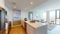 This contemporary high-rise residence is defined by clean lines, a neutral palette, and an abundance of natural light. The open-concept layout connects the kitchen and living area seamlessly, creating an inviting and functional space. The kitchen is outfitted with sleek flat-front cabinetry, a large center island with bar seating, quartz countertops, and a full suite of stainless steel appliances, including a French door refrigerator, microwave, and dishwasher. Warm hardwood flooring runs throughout, adding a natural contrast to the crisp white finishes. The living area is anchored by expansive floor-to-ceiling windows that frame breathtaking panoramic views of the surrounding cityscape and distant mountains, flooding the space with natural light. Both bedrooms continue the modern aesthetic, featuring the same sweeping views, hardwood and plank flooring, and ample natural light. The bathroom is finished with neutral tile, a soaking tub with a frameless glass sliding enclosure, a vanity with warm-toned countertops, and backlit mirrors. Recessed lighting throughout the residence ensures a bright, polished atmosphere at any hour.