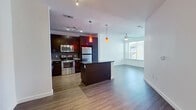 This thoughtfully designed 1-bedroom, 1-bathroom residence maximizes its single-floor layout with a contemporary open-concept living space. The combined kitchen and living area forms the heart of the home, anchored by a sleek kitchen island with dark granite countertops and bold pendant lighting. The kitchen is equipped with stainless steel appliances—including a range, over-the-range microwave, and refrigerator—paired with rich dark-toned cabinetry and a mosaic tile backsplash. Wood-style flooring runs throughout the main living areas, lending a warm, cohesive aesthetic. The living area is bright and open, benefiting from a large window that invites natural light and provides outdoor views, complemented by a ceiling fan overhead. A hallway connects the main living space to the private bedroom, which features plush carpeting, a ceiling fan, and a window framing greenery outside. The bathroom is clean and functional, offering a tiled soaking tub and shower combination, a dark-cabinet vanity with granite countertops, a large mirror, and matching wood-style flooring. White walls throughout the unit create a neutral, modern canvas well-suited for a variety of interior styles.