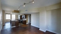 This single-floor space showcases a thoughtfully designed open-concept layout that seamlessly connects the kitchen and living areas. The kitchen is outfitted with rich dark-wood cabinetry, stainless steel appliances including a built-in microwave and range, and a central island that adds both functionality and visual appeal. The polished concrete floors throughout lend an industrial-modern character to the space, offering a striking contrast against the warm tones of the cabinetry and neutral-painted walls. Track lighting runs along the ceiling, providing adjustable, focused illumination across the open floor plan and enhancing the contemporary aesthetic. The living area offers a generous, versatile footprint ideal for a variety of furniture arrangements. A doorway visible from the main area leads to additional spaces, suggesting a well-organized floor plan. The ceiling height adds a sense of openness, while the clean white baseboards and smooth walls maintain a crisp, polished finish throughout. Natural light filters in from windows near the kitchen area, complementing the overhead lighting to create a bright and welcoming atmosphere. Overall, the space combines modern design elements with practical features for comfortable everyday living.