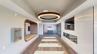 Located in Scottsdale, Arizona, this thoughtfully designed fitness center spans a generous single-floor layout, offering a comprehensive and upscale exercise environment. The space is divided into distinct functional zones, each finished with careful attention to detail and modern aesthetics.

The yoga and movement studio is a standout feature, boasting wide-plank hardwood-style flooring, a dramatic circular pendant light fixture with warm ambient glow, a sleek wall-mounted flat-screen TV, and a linear gas fireplace set against a white marble-look wall. A built-in curved wooden nook with cushioned seating adds a warm, organic architectural element, while floor-to-ceiling windows at the far end flood the room with natural light and outdoor views.

The main cardio floor is expansive and bright, lined with large panoramic windows overlooking the Scottsdale surroundings. It accommodates an array of equipment including treadmills, spin bikes, ellipticals, and stair climbers, all arranged across a durable combination of hardwood and multi-toned carpet tile flooring. A distinctive curved white reception-style console sits centrally, complemented by pendant lighting above.

A glass-walled strength room provides a dedicated free weights area with mirrored walls, dumbbell racks, and benches, offering both privacy and visibility. Throughout, the facility maintains a clean, contemporary palette of whites, warm woods, and soft neutrals, creating an inviting and motivating atmosphere.