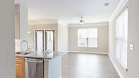Located at 4407 Hopson Rd in Durham, NC, this single-floor, one-bedroom, one-bathroom residence offers a thoughtfully designed layout that maximizes comfort and functionality. The open-concept living area seamlessly integrates the dining room, kitchen, and living room, creating an inviting, airy atmosphere bathed in natural light from multiple windows. Light-toned wood-look flooring flows throughout the main living spaces, complemented by crown molding and neutral wall tones that lend a clean, modern aesthetic. A ceiling fan anchors the living room, which opens to a private balcony via French doors. The kitchen is well-equipped with warm wood cabinetry, granite countertops, a white subway tile backsplash, stainless steel appliances including a refrigerator, range, and microwave, and track lighting overhead. The spacious primary bedroom features plush carpeting, a ceiling fan, and generous natural light. It provides direct access to a full bathroom appointed with white cabinetry, dual granite-topped vanities, and wood-look tile flooring, as well as a walk-in closet with wire shelving. A dedicated laundry room with washer and dryer hookups and an additional utility/storage area round out this efficient, move-in-ready residence.
