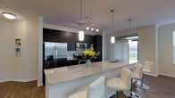 This single-floor space showcases a contemporary kitchen designed with a strong contrast between dark espresso cabinetry and light granite countertops. The kitchen is anchored by an expansive peninsula that doubles as a breakfast bar, accommodating multiple white upholstered bar stools for casual dining and entertaining. The granite surface extends generously, providing ample workspace throughout.

Stainless steel appliances, including a French door refrigerator with an in-door water and ice dispenser, a built-in microwave, and an electric range, are seamlessly integrated into the dark cabinetry for a cohesive, polished look. A white subway tile backsplash adds a clean, classic contrast to the bold cabinetry.

Lighting is a notable feature, combining a modern track lighting system above the cooking area with elegant pendant lights suspended over the peninsula, creating layered and functional illumination. The kitchen opens toward a naturally lit area with large windows dressed in sheer vertical blinds, allowing soft outdoor light to filter into the space. A decorative vase with fresh yellow flowers adds a warm, inviting touch to the overall modern aesthetic.