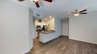 This single-floor residence offers a thoughtfully designed open-concept layout that seamlessly connects the kitchen and living areas, creating a bright and functional living environment. The kitchen is a standout feature, showcasing crisp white cabinetry, dark countertops, and a full suite of stainless steel appliances including a refrigerator, range, and built-in microwave. A peninsula with an integrated sink provides additional workspace and serves as a natural divide between the kitchen and the main living area, making it ideal for both everyday use and casual entertaining. Rich, dark hardwood-style flooring runs throughout the space, lending warmth and a cohesive aesthetic to the interior. The walls are finished in a neutral light gray tone, enhancing the sense of openness and complementing the modern design palette. Crown molding adds a refined architectural detail along the ceilings, while ceiling fans in both the kitchen and living areas ensure year-round comfort. Recessed lighting in the kitchen provides focused illumination, and a private entry door is visible from the main living space, contributing to a practical and well-organized floor plan overall.