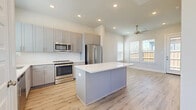 This well-designed single-floor residence offers a thoughtful layout across one bedroom, one bathroom, and a combined kitchen and living area connected by a hallway. The open-concept kitchen and living room serve as the heart of the home, featuring light wood-look flooring, recessed lighting, and a ceiling fan that contributes to a bright, airy atmosphere. The kitchen is outfitted with grey shaker-style cabinetry, a white subway tile backsplash, stainless steel appliances including a refrigerator, range, and microwave, and a central island with a crisp white countertop that provides additional workspace and seating potential. Multiple windows dressed with horizontal blinds allow generous natural light to fill the living area, which also benefits from outdoor views through a glass-paneled door. The bedroom is finished with plush carpeting, a ceiling fan, and windows that bring in abundant daylight. The bathroom features a grey vanity with granite-style countertops, gold-toned hardware, large-format tile flooring, and a glass-enclosed walk-in shower. Clean white walls and consistent design choices throughout the space create a cohesive, contemporary aesthetic.