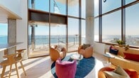 Located at 1515 Surf Ave in Brooklyn's iconic Coney Island neighborhood, this elevated single-floor living space offers a remarkable blend of contemporary architecture and breathtaking coastal scenery. The open-plan living room is anchored by soaring double-height floor-to-ceiling glass walls that flood the interior with abundant natural light while framing unobstructed panoramic views of the Atlantic Ocean, Coney Island Beach, and the surrounding Brooklyn skyline.

A wraparound outdoor terrace with warm hardwood decking and sleek glass railings extends the living space seamlessly to the outdoors, providing an exceptional vantage point over the beachfront below. The curved architectural detailing of the balcony above adds a distinctive, modern character to the building's exterior.

Inside, the living area features a built-in curved banquette seating arrangement set directly against the expansive windows, maximizing the ocean views. Light wood flooring, exposed concrete structural columns, and a textured brick accent wall contribute to the space's refined yet welcoming industrial-modern aesthetic. A bar counter area with warm wood cabinetry and richly paneled walls anchors one end of the room, adding both function and visual interest. The overall ambiance is bright, airy, and sophisticated, with the ever-present ocean horizon serving as a defining element throughout.