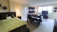 This single-floor studio space makes efficient and stylish use of an open floor plan, seamlessly integrating multiple functional zones within one cohesive layout. The sleeping area is anchored by a full-sized bed dressed in crisp green bedding, complemented by a dark wood headboard and a matching nightstand topped with a warm table lamp that adds a soft, inviting glow. A decorative beaded curtain serves as a creative visual divider, elegantly separating the sleeping zone from the rest of the space without the need for solid walls. Adjacent to it, a dark wood desk paired with a sleek ergonomic office chair establishes a dedicated workspace, while a modular open shelving unit displays decorative accents and personal items. The living area features a flat-screen television and comfortable seating, completing the multi-use layout. Natural light filters in through a window, brightening the space and complementing the neutral, warm-toned walls. Plush wall-to-wall carpeting runs throughout, contributing to the space's cozy and comfortable atmosphere. The overall aesthetic is modern and functional, with dark wood furniture providing a consistent design thread across all zones.
