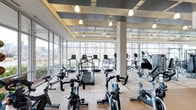 This single-floor fitness center is designed with both functionality and aesthetics in mind. The space is defined by expansive floor-to-ceiling windows along multiple walls, flooding the interior with abundant natural light while offering open views of the surrounding urban landscape — a feature that creates an energizing atmosphere for workouts.

The ceiling design is a notable architectural element, featuring a layered, coffered structure with inset lighting panels and pendant drop lights that provide consistent, well-distributed illumination throughout the space. Structural columns are integrated seamlessly into the layout without disrupting the open feel of the room.

The fitness floor is generously sized, accommodating a wide array of cardio equipment arranged in organized rows. Treadmills, elliptical trainers, stair climbers, and stationary cycling bikes are prominently featured, offering a comprehensive cardio training environment. The light-toned hardwood-style flooring complements the bright, airy interior and contributes to the clean, contemporary aesthetic.

Overall, the space presents a polished, modern fitness environment that balances high-end design with practical, well-organized equipment placement.