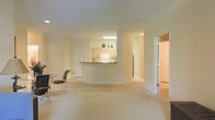 This single-floor residence is designed with an open-concept layout that creates a seamless flow between the living and kitchen areas. The main living space is finished with light-toned carpeting, contributing to a bright and airy atmosphere throughout. Neutral cream-colored walls and a white ceiling enhance the sense of openness and allow natural and artificial light to distribute evenly across the space.

A curved peninsula-style kitchen counter serves as the central focal point, separating the kitchen from the living area while maintaining visual connectivity between the two spaces. The kitchen features light wood cabinetry, a dark countertop surface, white appliances, and a sink positioned beneath an overhead fluorescent light fixture. Decorative items are displayed atop the upper cabinetry, adding a touch of personality to the space.

A bistro-style table and chairs are positioned in the living area near the kitchen, providing an informal dining option. Beyond the main living area, additional rooms are accessible through doorways visible in the background, suggesting further functional spaces within the residence. The overall ambiance is clean, minimal, and well-lit.
