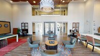 Located at 4650 N Central Ave in Phoenix, Arizona, this single-story community clubhouse delivers an impressive blend of contemporary design and social amenities. The grand entrance lobby immediately commands attention with its soaring double-height ceilings, a warm slatted wood ceiling detail, and a striking oversized pendant light fixture that anchors the space. Expansive floor-to-ceiling glass doors and windows flood the interior with natural light while providing views of the surrounding outdoor areas. The main lounge area features stylish mid-century modern seating arranged around a sculptural coffee table, accented by a bold patterned feature wall and a sleek bar-height counter. Wood-look plank flooring runs throughout, adding warmth and cohesion across the space. The game room area is well-appointed with a full-size pool table set on a vibrant red area rug, a dartboard wall, wall-mounted flat-screen televisions, a curated selection of board games, and dedicated high-top seating nooks. Colorful striped drapery panels frame the large windows overlooking the outdoor amenity area. The overall ambiance is energetic yet inviting, combining eclectic artwork, layered textures, and thoughtful furnishings to create a dynamic gathering space suited for both casual relaxation and lively recreation.