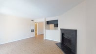This single-floor unit presents a practical and cohesive layout unified by warm parquet flooring that extends throughout every room. The living area serves as the centerpiece of the space, highlighted by a striking black brick fireplace with a raised hearth, adding a classic focal point to the otherwise clean, neutral interior. A textured popcorn ceiling and white walls create a bright, open atmosphere. A half-wall partition with dark upper cabinetry separates the living area from the kitchen, offering a pass-through design that maintains an open connection between the two spaces. The kitchen is efficiently arranged with dark cabinetry, white laminate countertops, and white appliances including an electric range and refrigerator. The bedroom is well-proportioned, receiving natural light through a window fitted with blinds, and connects conveniently to the bathroom area. The bathroom features a vanity with dark cabinet base, a mirrored medicine cabinet, and a separate compartment housing a full-size soaking tub with a tiled shower surround and toilet. Throughout the unit, the consistent parquet flooring and neutral palette contribute to a cohesive, functional living environment.