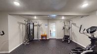Located at 6425 S Gessner Rd in Houston, Texas, this fitness center offers a dedicated, single-floor workout facility designed to support a well-rounded exercise routine. The space is finished with neutral gray walls, recessed lighting, and durable rubber flooring throughout, creating a clean and functional environment suited for regular physical activity.

The facility is divided into distinct workout zones. The cardio area features a Spirit treadmill and an elliptical machine, both positioned near a wall-mounted flat-screen television, allowing users to stay entertained during their workouts. A window with vertical blinds provides a source of natural light to this section of the room.

The strength training area is equipped with a multi-function cable machine and a power tower station that supports pull-ups, dips, and core exercises. A stationary bike rounds out the available equipment, offering additional low-impact cardiovascular options. A small utility table is conveniently placed within the space for personal items or cleaning supplies.

The facility is climate-controlled with visible HVAC vents throughout, and a security camera ensures a safe environment. A doorway connects to an adjacent area, contributing to ease of access. Overall, the fitness center presents a practical, well-maintained space for residents seeking convenient on-site exercise amenities.