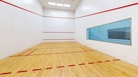 This dedicated racquetball court at 6005 State Bridge Rd in Johns Creek, Georgia, offers a well-maintained, regulation-size playing surface designed for competitive and recreational play. The court features a classic light maple hardwood floor with all standard red boundary lines and service markings clearly delineated throughout.

The enclosure rises to soaring ceilings, providing the vertical clearance required for full gameplay. The surrounding walls are finished in smooth, bright white panels — a standard construction that ensures consistent ball rebound — accented by bold red out-of-bounds lines running horizontally across all walls. Overhead, recessed and flush ceiling lighting delivers even, shadow-free illumination across the entire playing surface.

A standout architectural feature is the glass viewing wall and entry panel on one end of the court, allowing spectators to observe matches without interrupting play. This same wall also offers a sightline into an adjacent fitness area equipped with cardio equipment, suggesting that the court is part of a broader athletic or community facility. The overall environment is clean, functional, and purpose-built, making it an excellent venue for both casual players and serious competitors looking for a well-appointed indoor court.
