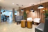 This single-floor amenity space offers a seamlessly integrated blend of wellness, fitness, and social areas, unified by a contemporary design language. The entrance greets visitors with exposed concrete columns, polished concrete floors, and a sputnik-style chandelier, setting an industrial-chic tone. Floor-to-ceiling glass doors invite natural light and street views into the welcoming lobby.

The central lounge corridor is a standout feature, with lush vertical greenery panels, large-scale textured wall art, and a curated mix of blue upholstered sofas, tufted seating, and warm pendant lighting. Slatted wood partitions and tall indoor plants create intimate seating zones while maintaining an open, airy flow throughout.

A social bar-height table area with wood-slat accent walls and integrated greenery niches provides a casual gathering spot. The fitness center is well-appointed with cardio equipment including treadmills and steppers, a rack of free weights, a wall-mounted flat-screen TV, and a Precor functional trainer with accessories such as a stability ball and exercise mat—all set beneath exposed concrete ceilings with linear LED lighting.

A glass-enclosed spa or wellness room features elegant frosted glass panels adorned with artistic ink-wash graphics, adding a serene, gallery-like quality to the space. Together, these distinct zones create a cohesive, resort-inspired amenity environment.