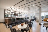 Located at 3 Hawkins Ave in Ronkonkoma, NY, this upscale amenity lounge offers a thoughtfully designed, open-concept floor spanning a generous single level. The space is defined by its contemporary aesthetic, blending light hardwood flooring, crisp white ceilings with linear beam detailing, and striking black steel-framed glass partitions that divide the floor into distinct yet visually connected zones.

The main lounge area features multiple seating arrangements to accommodate a variety of uses, including a bold mustard-yellow sectional sofa positioned beneath a multi-arm pendant light and flanked by large windows that fill the space with abundant natural light and outdoor views. Additional seating vignettes offer intimate conversation areas with stylish accent chairs and round tables throughout.

A sleek bar runs along one side of the space, lined with dark green upholstered barstools and illuminated by globe pendant fixtures, complemented by a bold geometric tile floor detail. A private conference room, enclosed by glass partitions, serves as a standout feature — anchored by a dark round table, upholstered chairs, and a dramatic sculptural gold chandelier that casts intricate shadow patterns across the grey walls. A separate lounge alcove with a channeled leather sofa and curated artwork adds a refined, gallery-like touch to the overall ambiance.