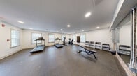 Located at 308 Scott St SW in Atlanta, Georgia, this fitness center offers a well-equipped exercise space designed to support a comprehensive workout routine. The room features durable rubber flooring throughout, providing a safe and functional surface for a variety of exercises. Recessed lighting illuminates the space evenly, complemented by multiple windows fitted with blinds that allow natural light to filter in while maintaining privacy.

The cardio section is equipped with multiple treadmills and an elliptical trainer, along with a stationary recumbent bike, all from the Spirit brand. A dedicated strength area includes a full dumbbell rack with a wide range of free weights, an adjustable weight bench, and additional resistance equipment. A wall-mounted flat-screen TV provides entertainment during workouts.

A standout architectural detail is the large wall mirror framed by exposed brick columns, which adds a striking visual element while expanding the perceived size of the room. Additional seating along one wall offers a convenient rest area. The space also includes a wall-mounted fire safety station and clearly marked emergency exit, ensuring safety compliance. The overall ambiance is clean, bright, and functional, making it a practical fitness facility for daily use.