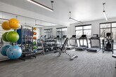 This modern fitness center offers a thoughtfully designed workout environment across a single open floor. The space is defined by a clean, contemporary aesthetic, featuring neutral-toned carpet flooring, crisp white walls, and a striking textured accent wall with a subtle wave-like pattern that adds visual depth to the interior. Linear pendant and recessed lighting fixtures illuminate the space evenly, contributing to an energetic yet comfortable atmosphere. Large windows along one wall invite abundant natural light and provide outdoor views, enhancing the open feel of the gym. Full-length mirrors are strategically installed along one side of the room, visually expanding the space while allowing users to monitor their form during workouts. The fitness center is comprehensively equipped with a row of treadmills and elliptical machines for cardio training, alongside a diverse selection of Hoist-brand strength and cable machines for targeted resistance work. A dedicated free weights area features a full dumbbell rack, an adjustable bench, and a colorful array of exercise balls and medicine balls on a storage rack, supporting a wide range of functional training activities. Flat-screen televisions are mounted on the walls for entertainment. The space is accessed through sleek, dark-framed glass double doors, reinforcing the facility's polished, upscale character.