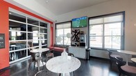 Located at 55 E 9th Ave in Columbus, Ohio, this single-floor space presents a sleek, modern aesthetic designed for comfort and functionality. The interior is characterized by clean lines, a neutral color palette punctuated by bold red accent walls, and large windows fitted with roller shades that allow generous natural light to fill the room while maintaining privacy.

The seating area features marble-top round tables paired with industrial-style metal chairs, creating a stylish yet practical gathering space. Decorative white floral arrangements add subtle elegance to the tables. A plush bench with red upholstery provides additional seating along one wall, complementing the room's color scheme.

A wall-mounted flat-screen television serves as a focal point in one area of the room, positioned above a sleek white credenza that houses what appears to be a beverage station, offering convenience for occupants. Abstract wall art adds a curated, contemporary touch nearby.

The dark tile flooring grounds the space and contrasts effectively with the bright white walls and furniture. Floor-to-ceiling glass partitions on one side introduce transparency and an open feel, connecting visual sightlines across the space. Overall, the room exudes a polished, modern ambiance suited for socializing or relaxing.