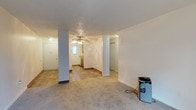 This single-floor space presents an open-concept layout that connects the main living area to the kitchen and additional rooms beyond. The living area is finished with wall-to-wall carpeting, lending a comfortable, understated feel underfoot. Two structural columns are positioned within the open space, defining subtle zones without fully enclosing them, and contributing to the architectural character of the interior.

A ceiling fan with a light fixture is centrally mounted on the textured popcorn ceiling, providing both airflow and ambient lighting to the main living zone. The walls are painted in a neutral off-white tone throughout, creating a clean, versatile backdrop.

Toward the rear of the space, the flooring transitions to hard surface material as it approaches the kitchen area, where warm wood-toned cabinetry is visible. A window in the kitchen allows natural light to filter into the interior. A doorway to the right leads to additional rooms, including what appears to be a closet with bi-fold doors. The front entry door is positioned to the left, providing straightforward access to the main living area. Overall, the space offers a practical, open arrangement suited for everyday living.