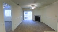 The living area of this single-floor space is thoughtfully designed to balance comfort and functionality. The room is carpeted throughout, providing a warm and soft underfoot feel that contributes to a cozy atmosphere. A ceiling fan with an integrated light fixture is centrally mounted, ensuring both air circulation and ambient lighting across the space.

A standout feature of the room is the tile-surround fireplace positioned along one wall, serving as a natural focal point and adding a classic, inviting element to the overall aesthetic. The fireplace surround features clean, neutral-toned tiling that complements the light-colored walls.

Natural light enters generously through a large sliding glass door, which opens to an outdoor balcony framed by white railings. This feature not only brightens the interior but also provides a seamless connection to the outdoors. The walls are finished in a neutral cream tone, offering a versatile backdrop suitable for a variety of décor styles. A textured ceiling and visible HVAC vent complete the practical details of this well-appointed living space.
