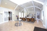 This single-story space presents a polished, welcoming environment well-suited for professional or administrative use. The central lobby serves as the heart of the layout, featuring warm beige tile flooring set in a diagonal pattern, soaring vaulted ceilings with exposed white beams, and recessed lighting that enhances the open, airy atmosphere. A decorative wrought-iron glass-top table anchors the space, complemented by built-in niche displays along the walls. Natural light streams in through multiple windows fitted with white plantation shutters, and French doors with louvered panels provide an elegant entry point.

Adjacent to the lobby, a reception area features a wainscoted service counter with wall sconces and decorative accents, offering a functional yet refined welcome space. A private office is accessed from the main area and includes a substantial executive desk, wicker seating for guests, plush carpeting, arched windows, and a built-in white cabinetry wall with decorative niches. A separate seating lounge with additional wicker chairs provides a comfortable waiting area. Throughout, the warm neutral palette, crown molding, and consistent architectural detailing create a cohesive and professional ambiance.