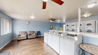 Located at 4277 Larson St in Olivehurst, CA, this community laundry facility offers a clean, comfortable, and functional environment for residents. The single-floor space is finished with durable wood-look flooring and light blue walls that lend a calm, welcoming ambiance throughout.

The main laundry area is well-appointed with a row of front-load washing machines centrally positioned for easy access, complemented by a bank of stacked dryers along the back wall. A built-in folding counter runs along one wall, providing ample surface space for sorting and folding laundry. Informational postings and signage are conveniently displayed near the machines.

A designated seating area features two patterned armchairs and an additional side chair beside large windows that allow natural light to fill the space, making the wait comfortable. Multiple ceiling fans are installed throughout, ensuring good air circulation.

The facility is accessed through a set of glass-paneled double doors, which also allow views to the surrounding outdoor area. Additional windows further brighten the interior and offer outdoor sightlines. A security camera is mounted near the ceiling, and a wall-mounted speaker provides audio throughout the space. The overall layout is open and organized, prioritizing ease of use and resident comfort.