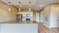 Located at 5050 Transit Rd in Depew, NY, this well-appointed one-bedroom, one-bathroom residence offers a thoughtfully designed single-floor layout with modern finishes throughout. The open-concept kitchen and living area serves as the heart of the home, anchored by a large peninsula with polished granite countertops and an integrated sink. White shaker-style cabinetry lines the kitchen walls, complemented by stainless steel appliances including a refrigerator, gas range, and built-in microwave. Three elegant pendant lights hang above the peninsula, adding warmth and visual interest to the space. Light wood-tone flooring runs throughout the main living areas, creating a clean and cohesive aesthetic. The bedroom is a comfortable, carpeted retreat featuring a window that brings in natural light and outdoor views of the surrounding landscape, along with access to a private balcony. The bathroom continues the home's refined design language with granite countertops and white cabinetry. A hallway connects the rooms efficiently, with light hardwood flooring maintaining a consistent feel. Neutral, warm-gray wall tones throughout the unit provide a calm, versatile backdrop suited to a variety of décor styles.