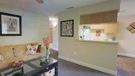 This single-floor residence offers a comfortable and functional layout designed for easy living. The living area is carpeted and finished in warm neutral tones, creating an inviting atmosphere. A dark-framed glass coffee table with wicker storage baskets anchors the seating area, which is complemented by a plush sofa adorned with decorative pillows and a vase of red floral accents. Wall décor, including a framed botanical art piece, adds character to the space. A pass-through window connects the living area to the kitchen, enhancing the open feel between the two spaces. The kitchen features light wood cabinetry, white appliances including a refrigerator, and a clean countertop workspace. The pass-through design allows natural sightlines across the unit, making the space feel more expansive. A hallway visible from the living room leads to additional areas of the residence, suggesting a well-organized floor plan. Textured ceilings and consistent neutral wall tones are carried throughout, contributing to a cohesive and understated aesthetic. Overall, the space balances practicality with a welcoming residential charm.