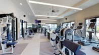 This fitness center offers a spacious, single-level layout designed to accommodate a comprehensive range of workout activities. The interior features a tray ceiling with recessed lighting and linear LED fixtures that provide consistent, even illumination throughout the space. A ceiling fan ensures comfortable air circulation during workouts.

The facility is generously equipped with a wide variety of exercise machines, including cardio equipment such as ellipticals and treadmills, as well as strength-training machines and a dedicated free weights section with a full dumbbell rack and mirrors lining the walls. A centrally positioned granite-topped counter provides a functional station within the workout floor.

Large windows along the perimeter allow abundant natural light to fill the room while offering views of the surrounding exterior. Striped, durable flooring adds visual interest and helps define distinct workout zones. A wall-mounted flat-screen TV enhances the experience for cardio users. The overall ambiance is clean, bright, and professional, creating an inviting environment suited for both casual and dedicated fitness routines.