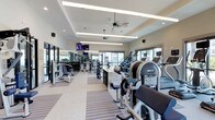 This fitness center offers a spacious, single-level layout designed to accommodate a comprehensive range of workout activities. The interior features a tray ceiling with recessed lighting and linear LED fixtures that provide consistent, even illumination throughout the space. A ceiling fan ensures comfortable air circulation during workouts.

The facility is generously equipped with a wide variety of exercise machines, including cardio equipment such as ellipticals and treadmills, as well as strength-training machines and a dedicated free weights section with a full dumbbell rack and mirrors lining the walls. A centrally positioned granite-topped counter provides a functional station within the workout floor.

Large windows along the perimeter allow abundant natural light to fill the room while offering views of the surrounding exterior. Striped, durable flooring adds visual interest and helps define distinct workout zones. A wall-mounted flat-screen TV enhances the experience for cardio users. The overall ambiance is clean, bright, and professional, creating an inviting environment suited for both casual and dedicated fitness routines.