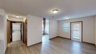 This single-floor residential unit offers a thoughtfully designed, open layout unified by light wood-style laminate flooring that flows seamlessly throughout every room. The living area is bright and welcoming, benefiting from a window with outdoor views and a door that opens to a private balcony, allowing natural light to fill the space. A separate bedroom is accessible via an open doorway, providing a clear division between living and sleeping areas.

The kitchen is well-appointed and functional, featuring rich medium-toned wood cabinetry spanning both upper and lower sections, white laminate countertops, and a double-basin stainless steel sink. White appliances include a range, microwave, dishwasher, and refrigerator, all arranged in a practical U-shaped configuration that maximizes workspace and storage.

The bathroom maintains the same consistent design language, with matching warm wood cabinetry beneath a single-basin vanity, white countertop, a large wall mirror with overhead lighting, and light wood-style flooring. Towel bars and a clean, neutral color palette contribute to a tidy and functional atmosphere.

Throughout the unit, white walls and flush-mount ceiling light fixtures create a clean, neutral backdrop. Interior doors feature warm wood frames consistent with the cabinetry, lending a cohesive aesthetic across all areas of the space.