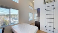 This contemporary single-floor space makes a strong first impression with its dramatic, soaring ceilings that create an expansive sense of volume and openness. The design is clean and minimalist, with crisp white walls that reflect natural light throughout the interior. Multiple large windows fitted with horizontal blinds line the walls at varying heights, flooding the space with daylight while offering views of the surrounding greenery and exterior environment.

The open layout seamlessly integrates the living area with a compact kitchenette tucked neatly into one corner. The kitchenette features dark espresso-toned cabinetry that provides a striking contrast against the light walls and flooring, complemented by a light-colored countertop and a built-in sink. The cabinetry extends vertically, making efficient use of the generous ceiling height.

Underfoot, light-toned hardwood-style flooring runs throughout the space, contributing to the bright and airy ambiance. Recessed ceiling lighting supplements the abundant natural light, ensuring the interior remains well-lit at all hours. The overall aesthetic is modern and understated, with a neutral color palette that offers a versatile and welcoming atmosphere. This space is well-suited for a variety of uses, combining functional design with an impressive architectural presence.