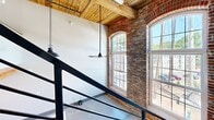 Located at 250 S Depot St in Pendleton, South Carolina, this two-story loft-style residence masterfully combines historic industrial architecture with contemporary design. The space is spread across two floors of nearly equal size, offering a thoughtful layout that maximizes every square foot.

The lower level features a striking open kitchen equipped with stainless steel appliances, light wood cabinetry, quartz countertops, and a central island. Concrete tile flooring grounds the space, while a bold black steel staircase ascends alongside exposed brick walls, adding an authentic industrial edge. Large arched windows on the main level flood the interior with abundant natural light and offer views of the surrounding neighborhood.

The upper level is anchored by the bedroom and hallways, continuing the loft aesthetic. Soaring ceilings with warm wood beam detailing and tongue-and-groove planking add rich texture overhead. The two bathrooms are distinctly finished: one features reclaimed hardwood floors, a marble-tiled tub/shower combo, and modern wood-toned vanity with black fixtures; the other showcases polished concrete floors and a sleek glass-enclosed marble shower. Together, they deliver a refined yet industrial sensibility consistent throughout the home. The overall ambiance is a compelling marriage of historic charm and modern sophistication.