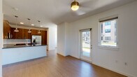 This single-floor residence offers a well-designed open-concept layout that seamlessly connects the living area to the kitchen. The living space features light wood-tone flooring, a ceiling fan with a brass-accented fixture, and a glass-panel exterior door that floods the area with natural light alongside a nearby window overlooking the surrounding neighborhood.

The kitchen is a standout feature, showcasing rich walnut-toned cabinetry that extends to the ceiling, a light-toned quartz island with an undermount sink and brass faucet, and a full suite of stainless steel appliances including a French door refrigerator, electric range, and built-in microwave. A subtle tile backsplash and pendant lighting with brass industrial-style fixtures complete the modern aesthetic.

The bedroom offers plush carpeting, a ceiling fan, and multiple windows providing generous natural light and open outdoor views of greenery and landscape. The bathroom features wood-look flooring, a soaking tub with large-format tile surround, a dark-toned vanity, and direct access to a walk-in closet with built-in shelving and hanging rods. Throughout, the space maintains a clean, contemporary ambiance with crisp white walls and cohesive modern finishes.