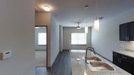 This single-floor space showcases a thoughtfully designed open-concept layout that integrates the kitchen and living areas into a cohesive, functional environment. The kitchen is equipped with a granite or stone-speckled countertop, a stainless steel double-basin sink with a contemporary arc faucet, and elegant pendant lights that hang overhead, adding both task lighting and visual interest. The flooring transitions from dark hardwood-style planks in the main living and kitchen areas to carpeting in the adjacent room, providing both durability and comfort throughout the space. Neutral gray walls create a clean, modern backdrop that complements the overall aesthetic. The living area benefits from natural light streaming through multiple windows fitted with horizontal blinds, contributing to a bright and airy atmosphere. A ceiling fan with a light fixture is centrally positioned in the living space, offering both airflow and ambient lighting. A doorway visible from the main area leads to a carpeted room, suggesting a private bedroom or secondary living space. The overall design is cohesive and contemporary, with a neutral palette and modern fixtures that give the space a polished, move-in-ready feel.