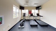 This single-floor fitness center is thoughtfully designed to support a comprehensive workout experience. The space is finished with durable rubber flooring throughout, warm cream-toned walls, and recessed panel lighting that creates a bright, energizing atmosphere. Large windows along multiple walls invite abundant natural light and offer views of the surrounding residential community, keeping the environment open and connected to the outdoors.

The cardio zone is equipped with Life Fitness treadmills, an elliptical trainer, and a rowing machine, all positioned beneath wall-mounted flat-screen televisions for entertainment during workouts. A dedicated strength training area features a multi-function cable machine paired with an adjustable workout bench, ideal for a wide range of resistance exercises. A full-length wall mirror in this section enhances the sense of space and allows for form monitoring during training.

The free weights area includes a well-stocked dumbbell rack with a range of weights neatly organized for easy access. Safety is prioritized with a clearly mounted fire extinguisher and AED unit on the wall. A direct exterior door provides convenient access to and from the surrounding outdoor areas, rounding out this practical and inviting fitness facility.