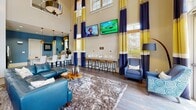 Located at 4600 Pear Grove Court in Glen Allen, Virginia, this single-story community clubhouse at Marshall Springs at Gayton Springs offers a well-appointed gathering space designed for resident amenity and social engagement. The centerpiece of the facility is a dramatic great room featuring soaring double-height ceilings adorned with bold navy, yellow, and white striped drapery that frame tall windows and French doors. These doors open directly to views of an outdoor pool area, flooding the space with natural light. Hardwood-style flooring anchors the main lounge, which is furnished with teal leather seating, plush area rugs, wall-mounted televisions, and a row of counter-height bar stools along a media ledge.

Adjacent to the lounge, a full kitchen provides stainless steel appliances, white cabinetry, granite countertops, and a mosaic tile backsplash set against rich blue-painted walls with light tile flooring. A dedicated café station nearby offers a self-serve coffee setup for residents. The billiards room features a coffered tray ceiling with ambient cove lighting, wall sconces, and a full-size pool table accompanied by comfortable lounge seating. A separate bar nook includes high-top tables, upholstered ottomans, and a beverage refrigerator beneath vibrant abstract wall art, completing this versatile amenity space.