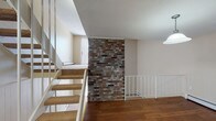 Located at 1 Juniper Dr in Yarmouth, Maine, this multi-level space spans three floors and offers a well-organized layout with a variety of functional living areas. A striking exposed brick accent wall serves as a central architectural feature, complementing the warm hardwood floors found on the main level. White railings line the staircase, connecting the floors with a clean, classic aesthetic.

The galley-style kitchen is outfitted with warm oak cabinetry, dark granite-look countertops, a stainless steel sink, and white appliances, including a refrigerator and dishwasher. Hardwood flooring carries through the kitchen, adding warmth and durability to the space.

The living area on the lower level features carpeted flooring, a brick fireplace surround, and baseboard heating. Sliding glass doors dressed with vertical blinds provide access to the outdoors and allow natural light to fill the room. Upper-level rooms continue the carpeted theme and feature vaulted ceilings, generous closet space — including a walk-in closet with wire shelving — and additional sliding door access. The bathroom includes a full tub and shower combination with wood-look flooring. Throughout all three levels, the neutral palette and thoughtful layout create a comfortable, move-in ready environment.