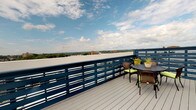 The Phase I Roof Deck is a multifaceted space combining a striking outdoor deck with thoughtfully designed interior common areas. The rooftop terrace is defined by its bold navy slatted railings and durable grey composite decking, creating a contemporary aesthetic. From this elevated vantage point, panoramic views stretch across the surrounding treetops, neighboring rooftops, and open sky, making it an ideal setting for outdoor seating and relaxation.

The interior spaces reflect a distinctive industrial-loft character, highlighted by soaring ceilings with exposed ductwork and a combination of polished concrete and warm hardwood flooring. Large, multi-pane windows flood the rooms with abundant natural light while framing expansive views of the surrounding urban and natural landscape. Wall sconces provide warm, ambient lighting throughout the lounge areas, which are furnished with comfortable sofas, accent chairs, and area rugs in rich, earthy tones.

The kitchen area features dark cabinetry, granite countertops, stainless steel appliances, and a peninsula with bar seating—combining functionality with style. Hallways with warm wood-toned doors connect the various areas seamlessly. Together, the indoor and outdoor elements create a versatile, inviting environment with a strong sense of openness and urban sophistication.