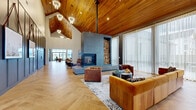 This expansive single-floor gathering space showcases a masterful blend of modern and rustic design elements. The centerpiece of the main lounge is a striking floor-to-ceiling concrete fireplace surround, flanked by a neatly stacked firewood display, creating a dramatic focal point beneath soaring vaulted ceilings clad in warm natural cedar. Elegant chandelier lighting complements the cathedral ceiling, while generous herringbone-patterned hardwood flooring flows seamlessly throughout the entire space.

The main seating area is anchored by a substantial sectional sofa in rich amber velvet, paired with tufted leather armchairs and layered area rugs, all oriented around the fireplace for an inviting atmosphere. Sheer floor-to-ceiling drapery lines the expansive windows and glass doors, diffusing abundant natural light and offering views to the outdoors.

A dedicated bar and kitchenette area features a sleek white quartz island with seating, pendant lighting, a textured tile backsplash, open brass shelving, and built-in appliances including a beverage cooler. Adjacent to this is a secondary lounge zone and a glass-enclosed dining room, providing versatile spaces for gathering or private meetings.

Dark charcoal board-and-batten wall paneling, curated artwork displayed on leather straps, and thoughtful architectural transitions between zones give the space a cohesive, upscale character throughout.
