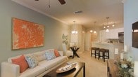 This single-story residence features an open-concept floor plan that connects the living, dining, and kitchen areas into one cohesive and functional space. The living room is anchored by a plush cream sofa adorned with blue and coral accent pillows, complemented by a dark wood coffee table. A bold orange abstract canvas adds a striking focal point to the soft sage-green walls, while crown molding lends a refined architectural touch. A ceiling fan provides both style and comfort overhead. The space flows naturally into a cozy sitting or dining nook, furnished with a classic X-base table and a cushioned accent chair, illuminated by a pendant chandelier. Adjacent to this is a well-appointed kitchen featuring white cabinetry, a bar-style counter with woven rattan stools, and stylish pendant lighting that enhances the warm ambiance. Arched architectural details frame transitions between spaces, adding character throughout. Neutral carpeted flooring runs throughout the main living areas, tying the coastal-inspired color palette of cream, coral, and sage together into a cohesive, welcoming atmosphere.