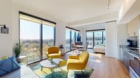 This contemporary single-floor unit is defined by its striking floor-to-ceiling windows, which flood the open-plan interior with natural light and frame panoramic elevated views of the surrounding treetops and urban skyline. The main living area is generously proportioned, featuring warm hardwood flooring, clean white walls, and a seamless flow between the lounge, dining, and sleeping zones. The lounge is anchored by bold yellow accent chairs and a colorful area rug, creating a vibrant yet sophisticated atmosphere. A compact dining nook with a round table and modern chairs sits adjacent, leading to a sleeping area that is thoughtfully separated yet open to the expansive views. Large roller shades offer adjustable privacy throughout. The kitchen is efficiently designed with two-tone cabinetry — light maple uppers paired with darker lower units — complemented by a dark countertop, a neutral tile backsplash, and a full suite of stainless steel appliances including a range, microwave, and dishwasher. The bathroom features marble-look tile flooring, a dark stone vanity countertop with warm wood cabinetry, and a backlit mirror, creating a clean and modern aesthetic. Throughout, the unit balances contemporary design with functional living, all bathed in abundant natural light.