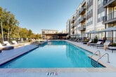 Located at 368 Sublime Summer Lane in Clover, South Carolina, this outdoor amenity space offers a resort-caliber pool experience within a modern residential community. The centerpiece is a generously sized swimming pool with crystal-clear blue water, complemented by a separate spa, both framed by a broad, light-toned concrete pool deck. Wicker chaise lounges with plush white cushions are arranged throughout the deck, providing ample space for sunbathing and relaxation. Shaded cabanas and market umbrellas offer respite from the sun, creating a range of comfortable seating options for residents. A section of well-maintained green turf adds a soft, natural contrast to the hardscape surroundings. The pool area is enclosed by sleek black iron fencing, beyond which a natural tree line of mature deciduous trees provides a verdant, private backdrop. The multi-story residential building rises alongside the pool courtyard, featuring contemporary architecture with large windows, private balconies with metal railings, and a neutral palette of gray and white siding. The overall ambiance is polished and resort-like, designed to encourage outdoor leisure and community gathering under open Carolina skies.