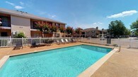 Located at 51 Northtown Dr in Jackson, Mississippi, this residential community offers a welcoming environment complemented by well-maintained outdoor amenities. The standout feature of the property is its generously sized outdoor swimming pool, which boasts crystal-clear blue water and a spacious surrounding deck. The pool area is thoughtfully appointed with lounge chairs and a shaded seating arrangement, providing residents with an ideal setting for relaxation and recreation under the Mississippi sun. A white perimeter fence encloses the pool area, ensuring both safety and a clean, classic aesthetic. Vibrant crepe myrtle trees in full bloom add a splash of color and natural beauty to the grounds, enhancing the overall curb appeal of the community. The multi-story residential buildings visible in the background feature a warm brick and neutral-toned exterior, set against a backdrop of mature trees and open sky. The single-floor layout of the space ensures ease of accessibility throughout. The combination of attractive landscaping, inviting communal spaces, and a well-kept exterior creates a comfortable and appealing living environment for residents in the heart of Jackson, MS.