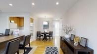 This single-floor residence offers a welcoming and well-appointed living environment characterized by clean lines and a light, airy atmosphere. The open layout allows natural light to flow freely throughout, complemented by recessed lighting and a stylish pendant fixture that anchors the central dining area. Warm hardwood floors extend across the main living and dining spaces, adding a sense of continuity and warmth to the interior.

The dining area features a round dark-wood table paired with tufted leather chairs, creating an intimate yet functional setting. A decorative area rug with a bold geometric pattern adds visual interest underfoot. Adjacent to the dining space, a sideboard displays tasteful décor, including a sculptural vase with branches and framed artwork, lending the room a curated, sophisticated feel.

The kitchen is accessible directly from the main living area and features warm wood cabinetry and essential appliances. A doorway leading to additional rooms is visible from the main space, hinting at a thoughtfully connected floor plan. The walls are finished in soft, neutral tones that enhance the brightness of the space and provide a versatile backdrop. Overall, the home presents a cohesive, comfortable environment that balances style and everyday functionality.