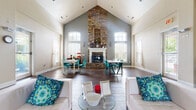 Located in Tinley Park, Columbus, Ohio, this single-floor community space makes a striking first impression with its dramatic vaulted ceilings that rise to a sharp peak, lending an expansive, airy quality to the interior. The centerpiece of the room is a floor-to-ceiling stacked stone fireplace framed by a crisp white mantel, anchoring the space with a warm, rustic elegance against a soft grey accent wall.

Rich hardwood-style flooring runs throughout, complementing the neutral warm-toned walls and white trim. Arched and multi-pane windows are positioned at varying heights, flooding the room with natural light and offering pleasant outdoor views. A grand arched entryway with double glass-paned doors provides a welcoming formal entrance, accented by a decorative chandelier overhead.

The open layout is thoughtfully arranged to serve multiple functions simultaneously. A comfortable lounge area with white modern sofas and vibrant teal accent pillows anchors one end, while multiple dining tables with bold teal chairs are positioned near the windows, ideal for gatherings or casual meetings. A wall-mounted television and water cooler near the secondary entrance add practical amenities. The overall ambiance is bright, modern, and inviting — a versatile space well-suited for community events and everyday use.
