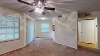 Located at Lifestyle Blvd in Palm Harbor, Florida, this single-story unit offers a practical and well-organized layout suited for comfortable everyday living. The living area is finished with wall-to-wall carpet and features a ceiling fan with integrated lighting, a textured ceiling, and a sliding glass door alongside a window that invites natural light into the space. Built-in shelving on a dividing half-wall adds a functional design element, while a pass-through connects the living area to the kitchen. The kitchen is equipped with black appliances including an electric range, over-the-range microwave, and a full-size refrigerator. Light wood cabinetry with dark countertops and a mosaic tile backsplash give the kitchen a clean, contrasting aesthetic, and hardwood-style flooring completes the space. The bedroom is generously proportioned with plush carpeting, a ceiling-mounted light fixture, a textured ceiling, and access to a dedicated closet with louvered doors offering ample storage. The bathroom is finished with hardwood-style flooring and includes a single-sink vanity with light wood cabinetry, a dark countertop, a framed mirror with overhead lighting, and a tub-shower combination. The overall layout is efficient and cohesive, making thoughtful use of the available space throughout.