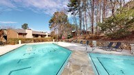 Located at 200 Ridgewood Dr in Raleigh, NC, this single-level outdoor amenity area is centered around a generously sized swimming pool bearing the distinctive Averett at North Hills branding on its floor. The pool is framed by a broad concrete deck that provides ample space for lounging and recreation. Lounge chairs and seating areas are arranged poolside, offering comfortable spots to relax and take in the natural surroundings.

The space is enveloped by a lush, wooded backdrop of tall pines, deciduous trees, and manicured shrubs, lending the area a serene, park-like atmosphere. A stone retaining wall runs along one side of the pool, adding a rustic, natural element to the overall design. Potted plantings and ornamental grasses accent the hardscape, contributing to the well-maintained landscaping.

Access to the pool area is provided through glass-panel entry doors set within a brick-accented building facade, which also features posted pool rules and signage. The surrounding buildings, visible in the background, reflect a mid-century architectural character with brick exteriors. Open to the sky, the space benefits from abundant natural light throughout the day, making it an inviting outdoor destination for residents of the community.